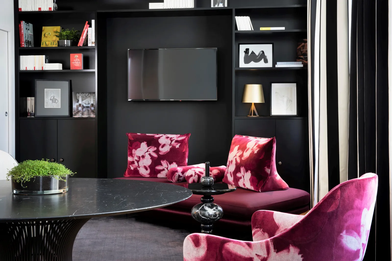 Modern lounge at Hôtel Cardinal Bordeaux featuring black walls, built-in shelving unit, TV screen, pink floral accent chairs, black marble round table, green plant and contemporary decor