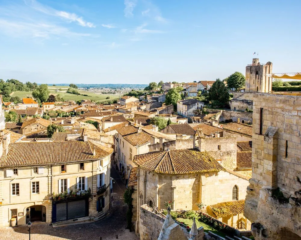 Panoramic view of a fortified French town featuring terracotta roofing, golden stone buildings, a Romanesque church, and watchtower, surrounded by green countryside.