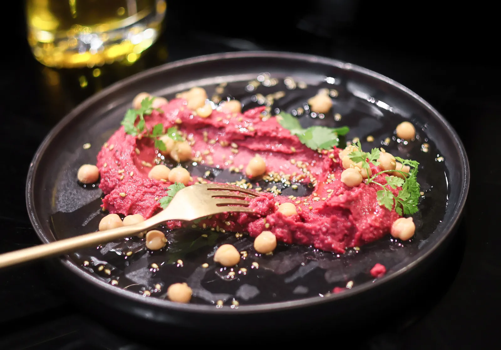 Black plate featuring a vibrant pink beetroot purée topped with golden chickpeas, microgreens, and a gold fork, with a glass of white wine in the background