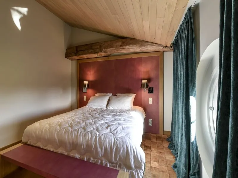 Elegant bedroom at Château Beauregard in Bordeaux featuring a queen-size bed with white linens, burgundy headboard, light wood ceiling with exposed beams, and green drapes.
