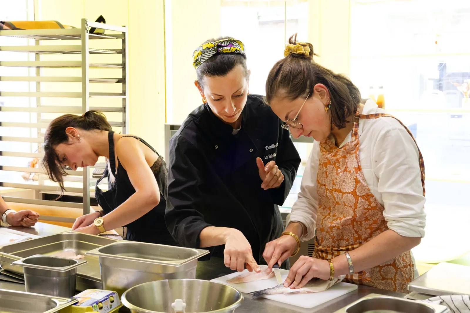 Three women in a professional kitchen in Bordeaux, working together on food preparation. A chef in a black jacket leads the demonstration while two other participants observe and assist.