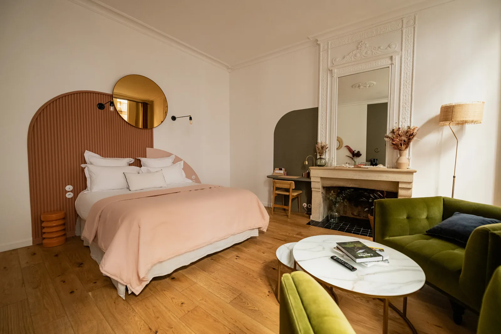 Elegant bedroom at La Maison Fernand in Bordeaux, featuring a rounded terracotta headboard, pale pink bedding, a gold circular mirror, a white fireplace, light wooden floors, and an olive green sofa lounge area