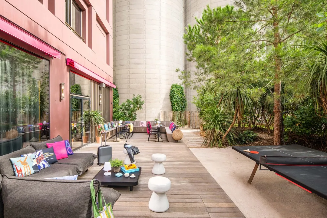 Outdoor terrace of Moxy Bordeaux featuring modern furniture, gray sofas, colorful cushions, trampoline, pink building facade and lush greenery in the background