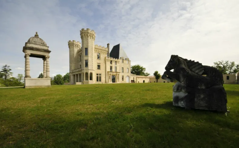 Old Lormont Castle in Bordeaux featuring crenellated stone towers surrounded by green lawns and contemporary sculptures under a partly cloudy sky