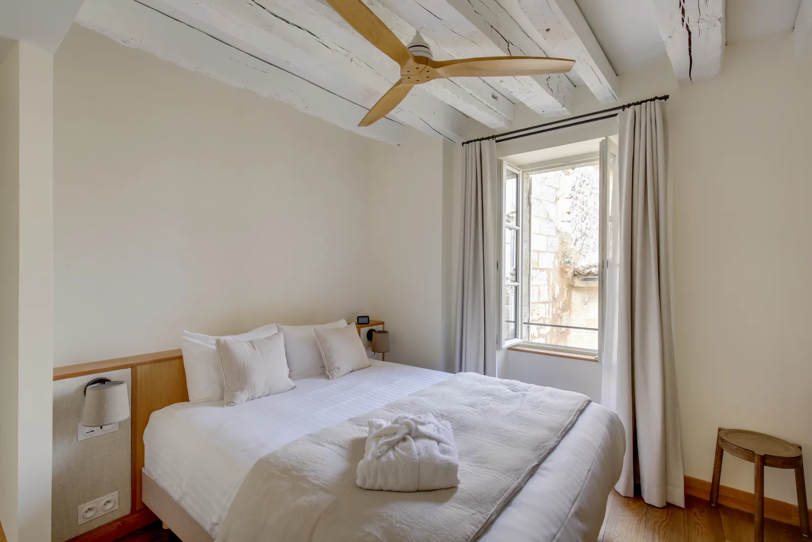 Bright bedroom at Badon Boutique Hotel in Bordeaux featuring a queen-size bed, white beam ceiling, wooden ceiling fan, French doors opening to a balcony, and minimalist light wood furniture.