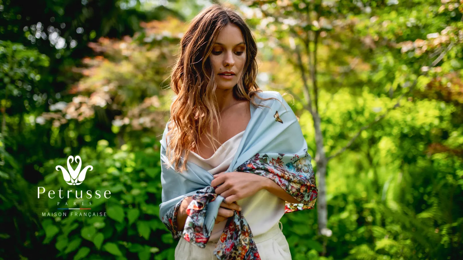 Woman wearing a light blue shawl with floral patterns in a lush garden, Petrusse French House collection