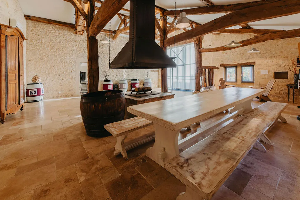 Rustic interior of Château Rol Valentin in Bordeaux featuring exposed wooden beams, white farmhouse table, central black fireplace and raw stone walls
