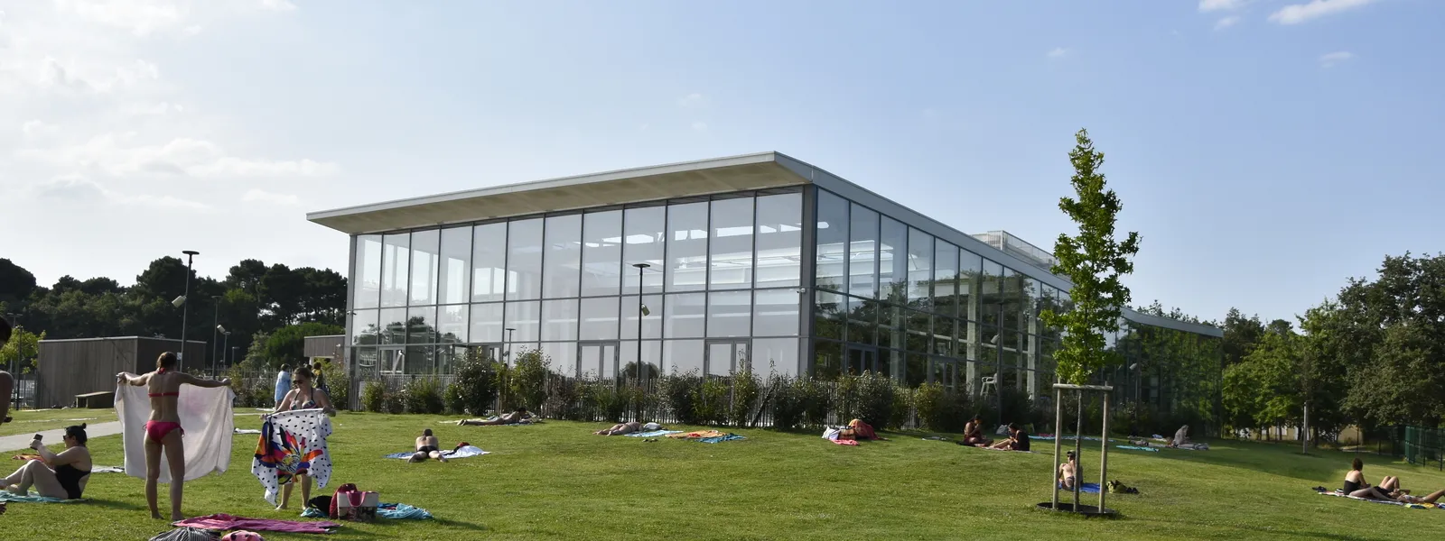 The Pinsan Swimming Pool in Eysines featuring a modern glass-fronted building surrounded by a green lawn where visitors relax and sunbathe on a sunny day.