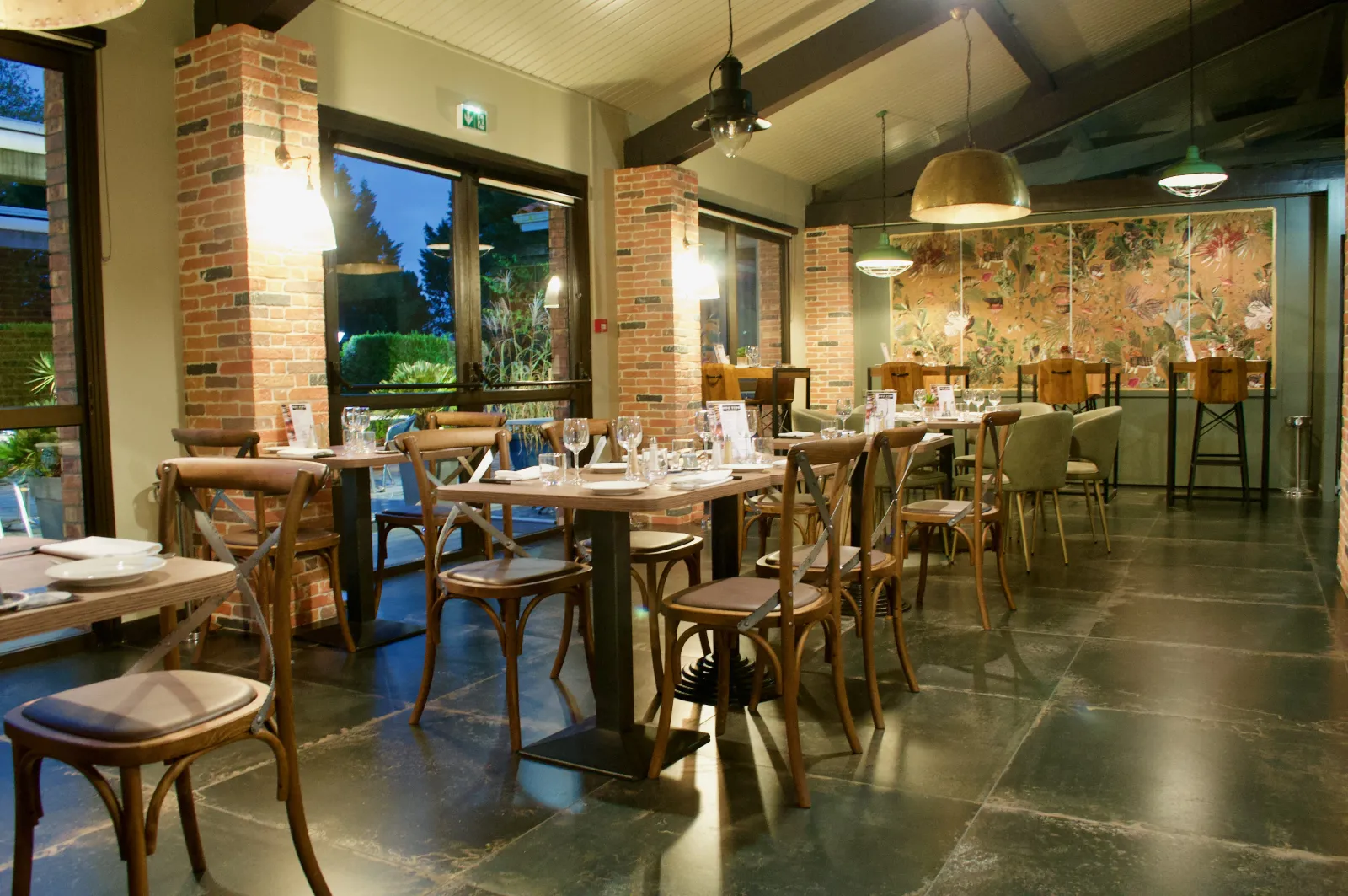 Elegant dining room with wooden tables, bistro chairs, exposed brick walls, large windows overlooking a lit garden, floral wall decoration and golden pendant lights.