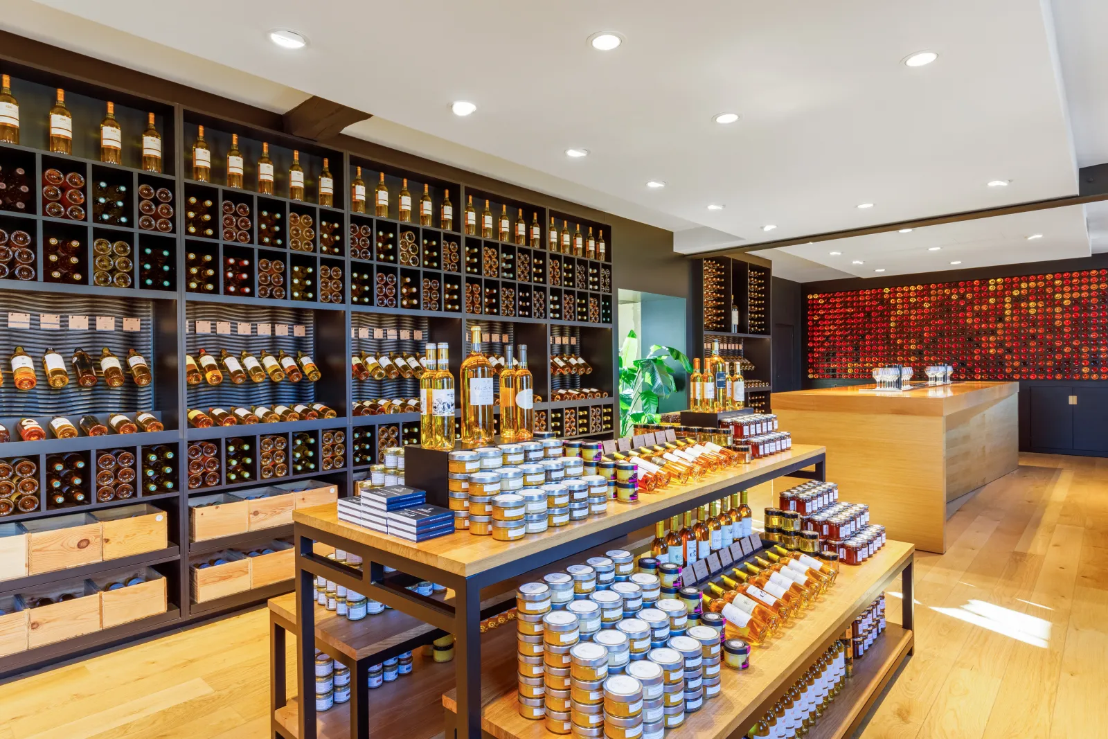 Elegant interior of the Maison du Sauternes in Bordeaux featuring black wall shelving displaying white wine bottles and regional products, light wood tasting tables filled with jars and bottles, a blonde wood reception counter, modern white ceiling with r