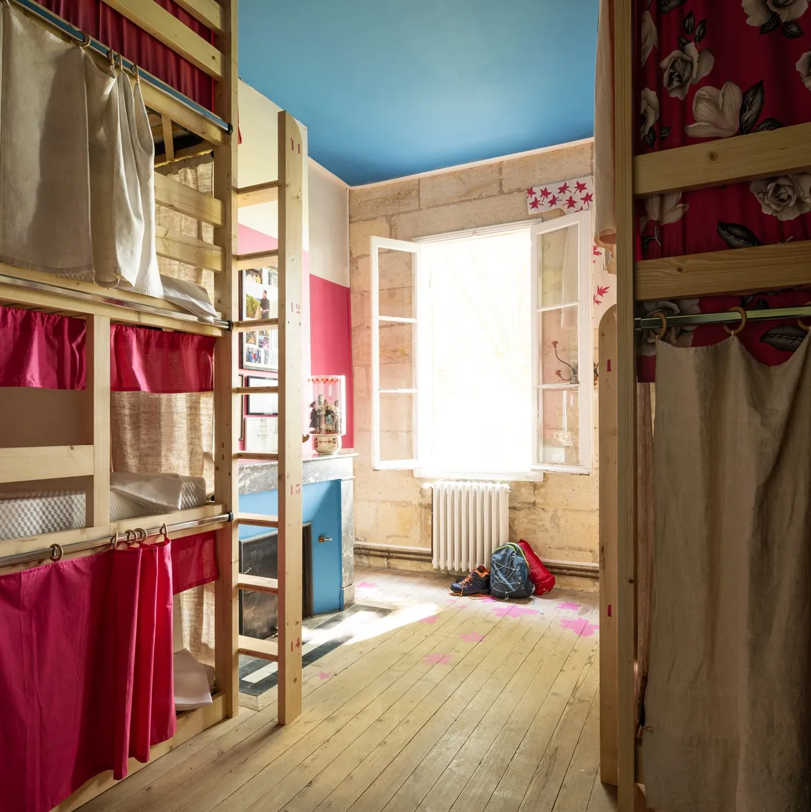 Joualles dormitory in Bordeaux: spacious bedroom with wooden bunk beds, red and beige curtains, bright window overlooking the street, blue ceiling, light wooden floor.