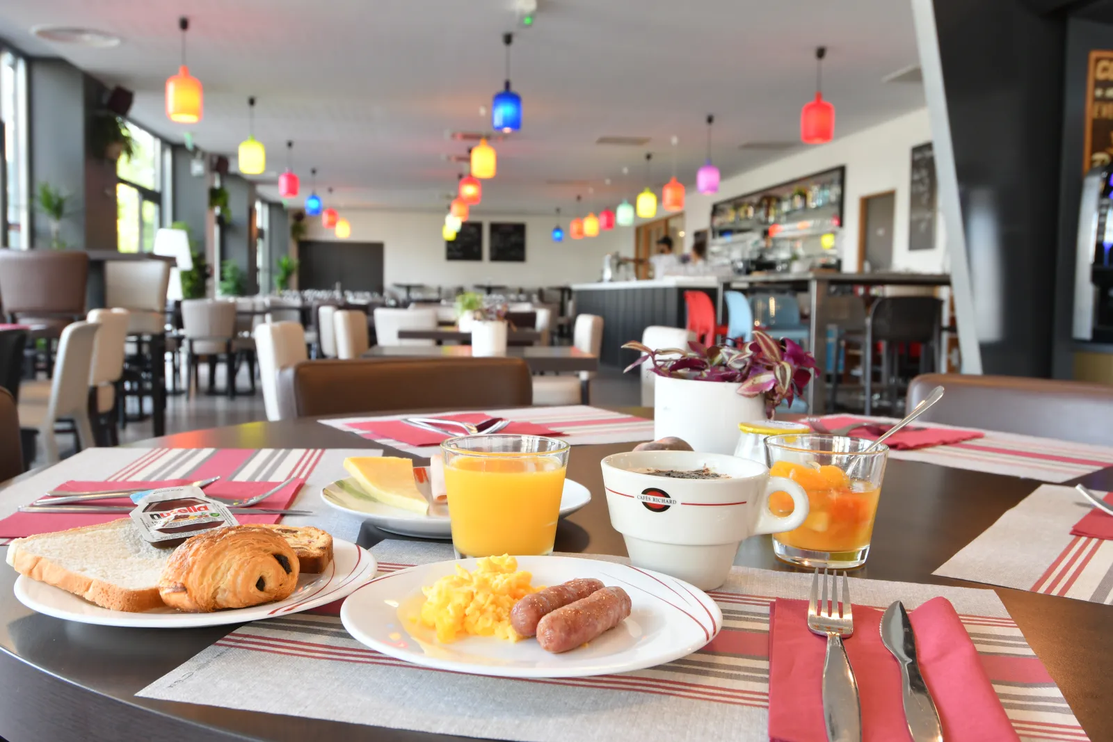 Vibrant breakfast room with set tables, pastries and fresh orange juice, featuring colorful pendant lamps and modern decor