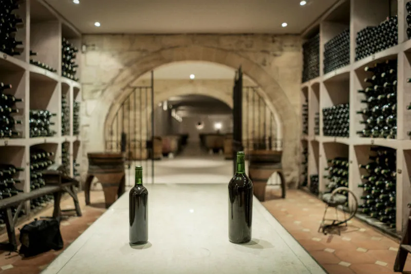 Wine cellar of Château Prieuré-Lichine in Bordeaux featuring two wine bottles in the foreground and storage racks filled with bottles on either side, with vaulted tunnels in the background