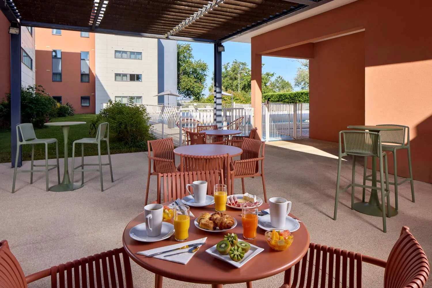 Covered terrace of a Bordeaux hotel featuring breakfast tables, red and green chairs, coffee and orange juice, residential buildings in the background under blue sky.