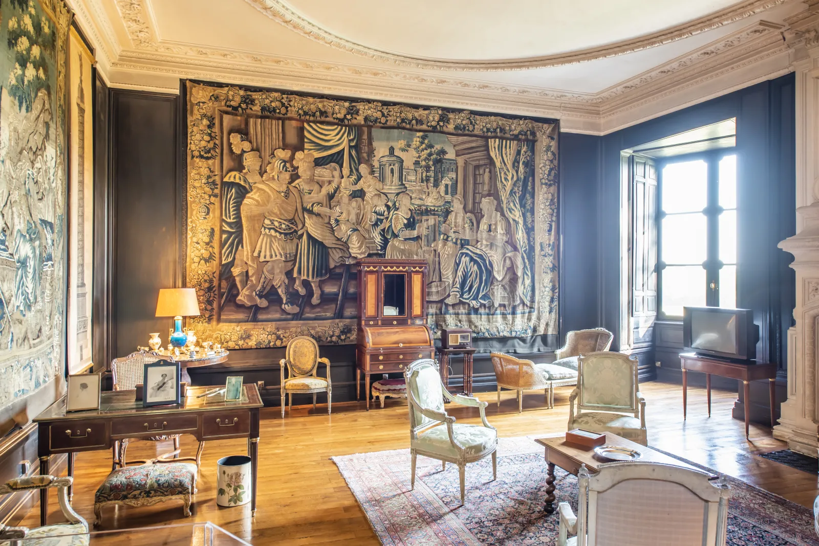 Elegant drawing room at Château de La Brède featuring period furniture, gilt tapestries, coffered ceiling, and tall windows overlooking the grounds.