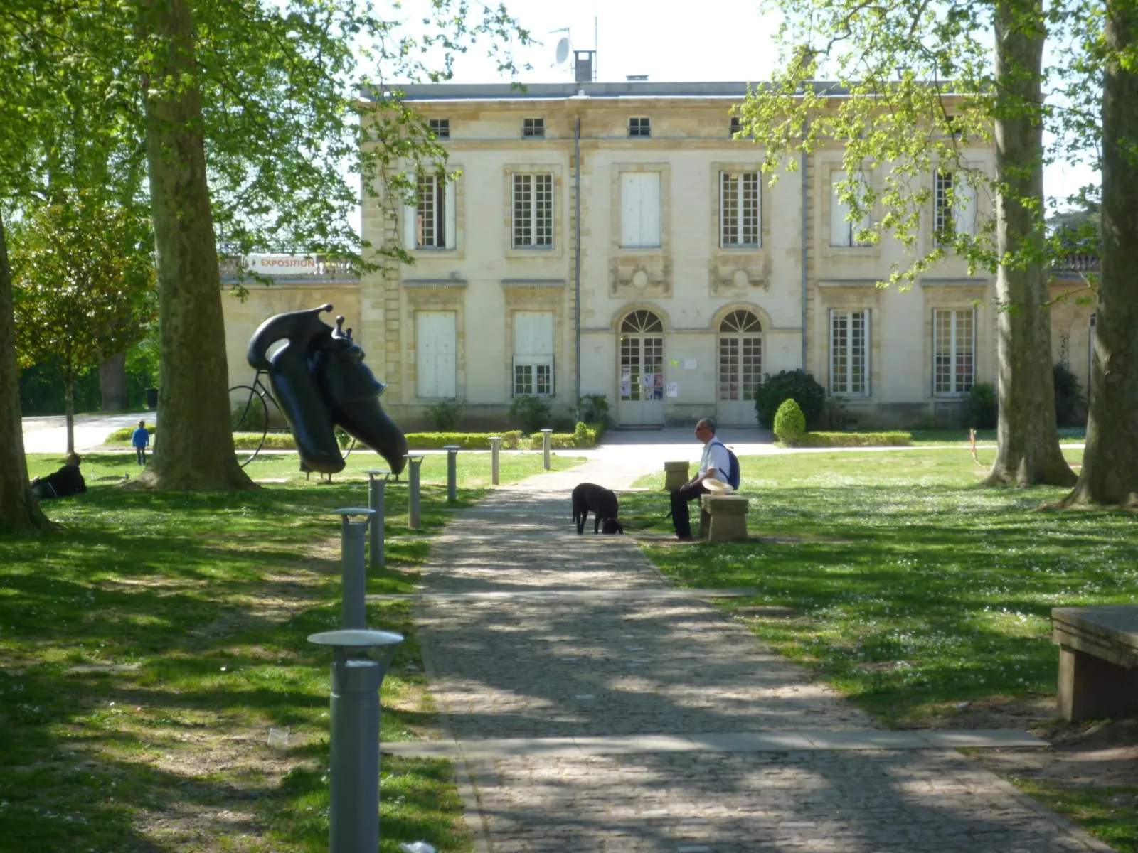 Palmer Park in Bordeaux featuring a classical mansion, abstract black sculpture, tree-lined pathway, and visitors relaxing on the lawn