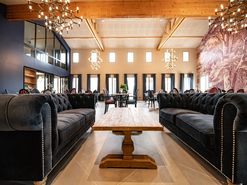 Elegant reception hall at Château Fage featuring two black Chesterfield sofas facing each other, wooden coffee table, exposed beam ceiling with contemporary chandeliers, and large windows overlooking Bordeaux vineyards.