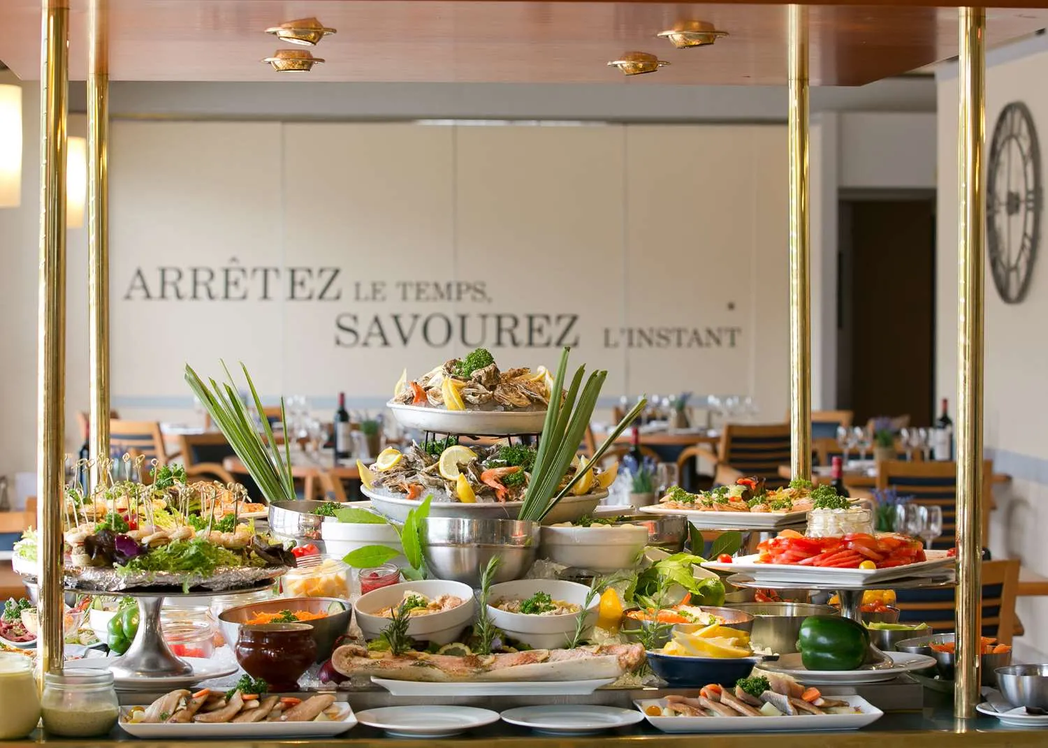 Gourmet buffet display featuring seafood platters, fresh salads, fish, and colorful vegetables in a restaurant overlooking Bordeaux. French motto on wall encouraging to savor the moment.