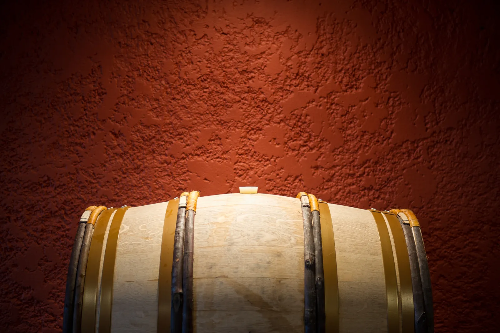 Wooden wine barrel with golden metal bands against a textured terracotta wall in the cellars of Château de Rayne Vigneau in Bordeaux.