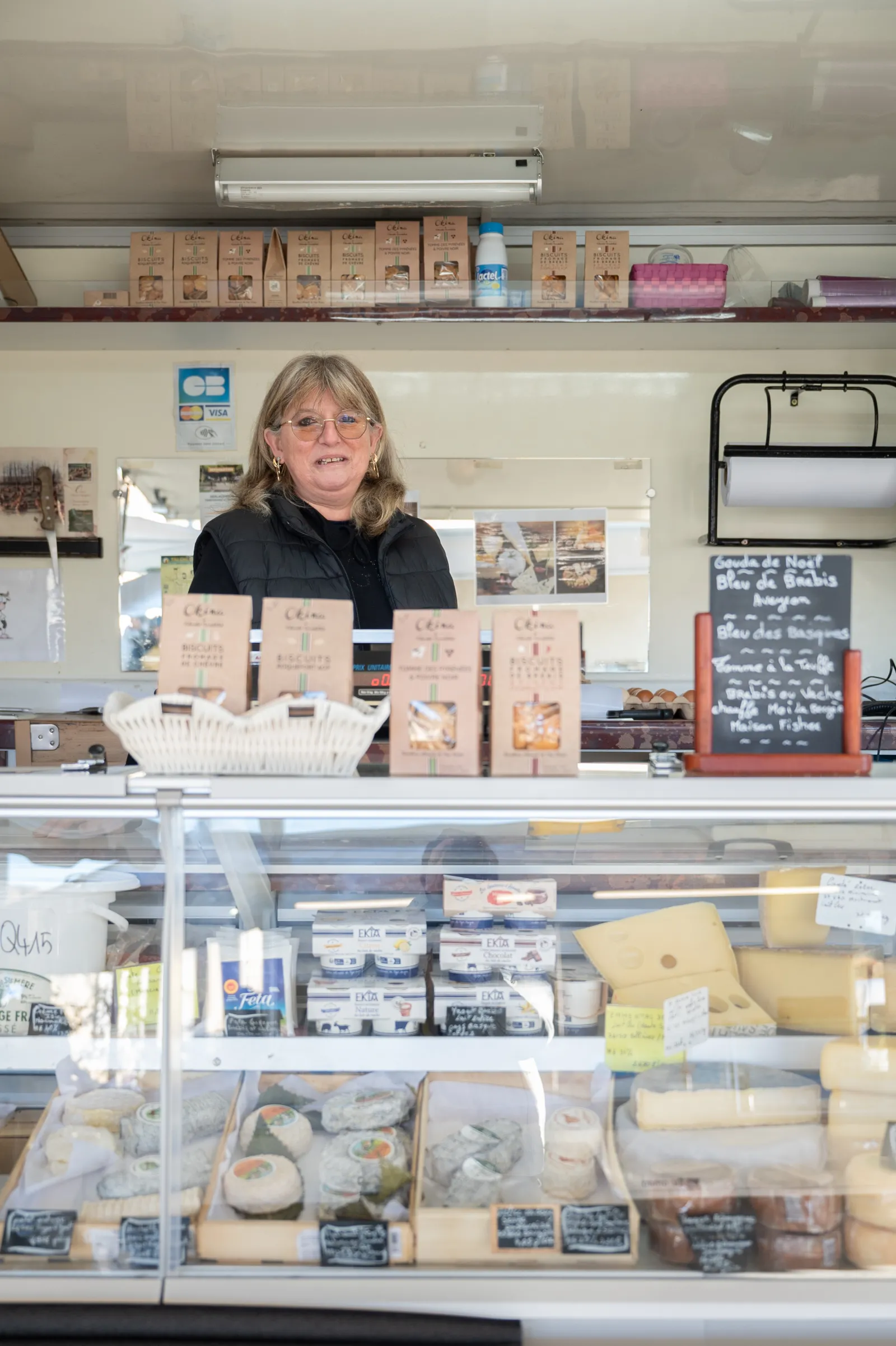 Smiling woman standing behind a counter at an artisanal cheese shop in Halle Vettiner, Bouliac, Bordeaux, with cheeses and dairy products displayed in a refrigerated showcase and on shelves.