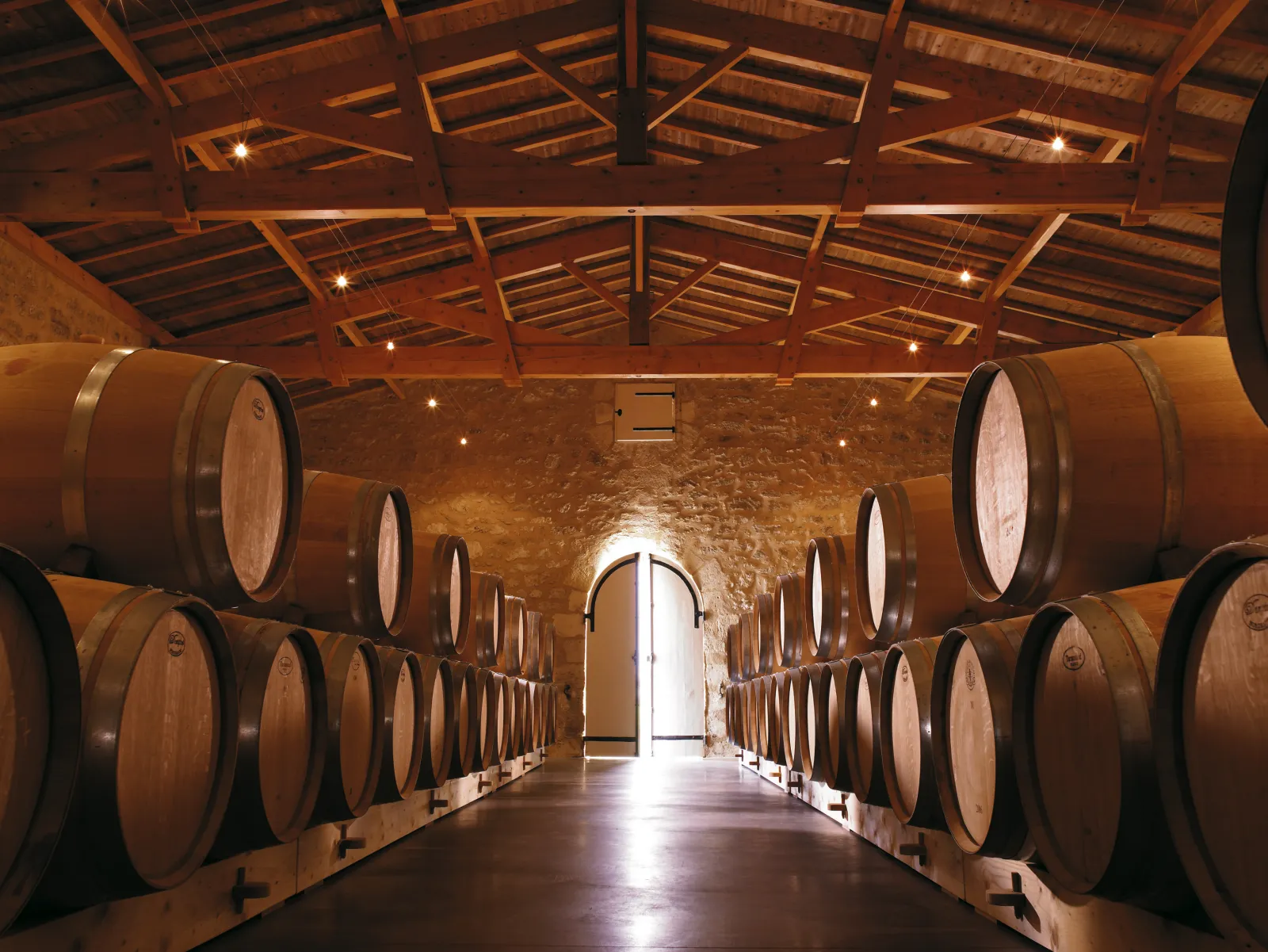 Winemaking cellar at Clos des Jacobins in Bordeaux featuring rows of oak barrels under a wooden beamed ceiling with open doors at the end.