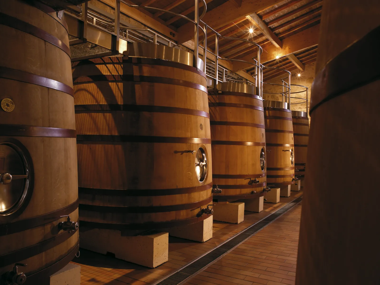 Row of oak barrels with black metal bands in a lit winery cellar, featuring metal catwalks and exposed beams.