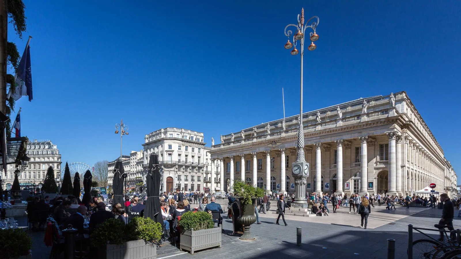 The Grand Theatre of Bordeaux, a neoclassical building with majestic white columns, ornamental façade and clock, overlooking a bustling public square under clear blue sky.