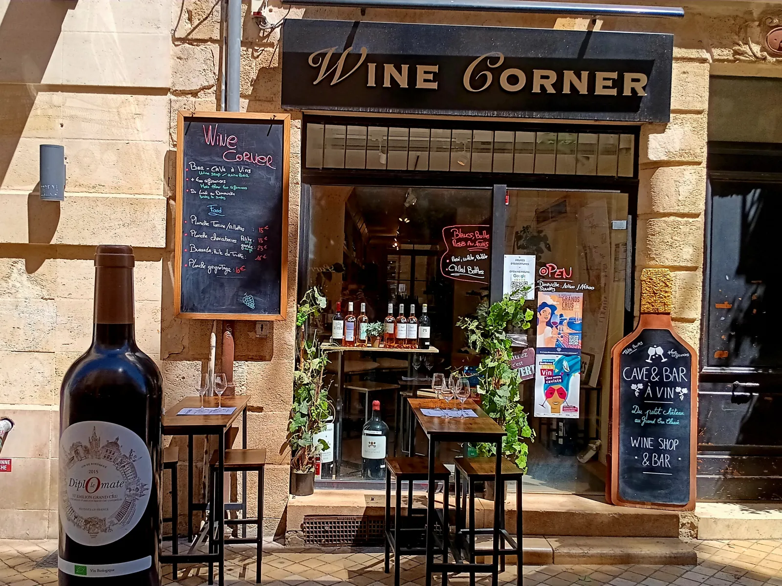 Storefront of Wine Corner, a wine shop and bar in Bordeaux, featuring bottle displays, high-top seating area, chalkboard menu, and wooden signage.