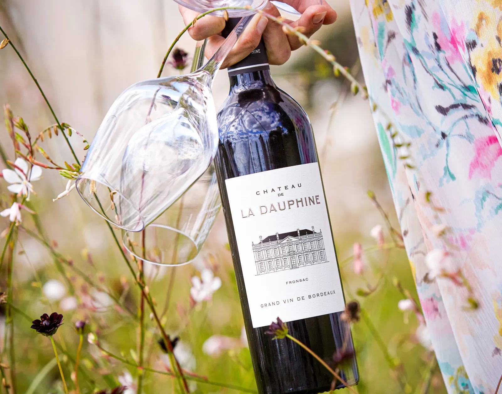 A bottle of Château de La Dauphine red wine from Fronsac, a grand Bordeaux wine, held with a wine glass surrounded by pink and white wildflowers.