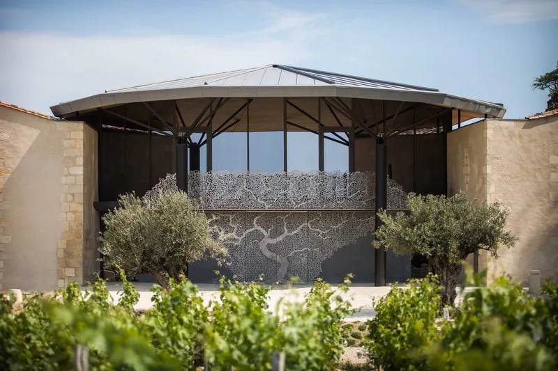 Modern tasting building with arched metal roof, glass facade and perforated panels, surrounded by vineyard rows at Château Kirwan in Bordeaux