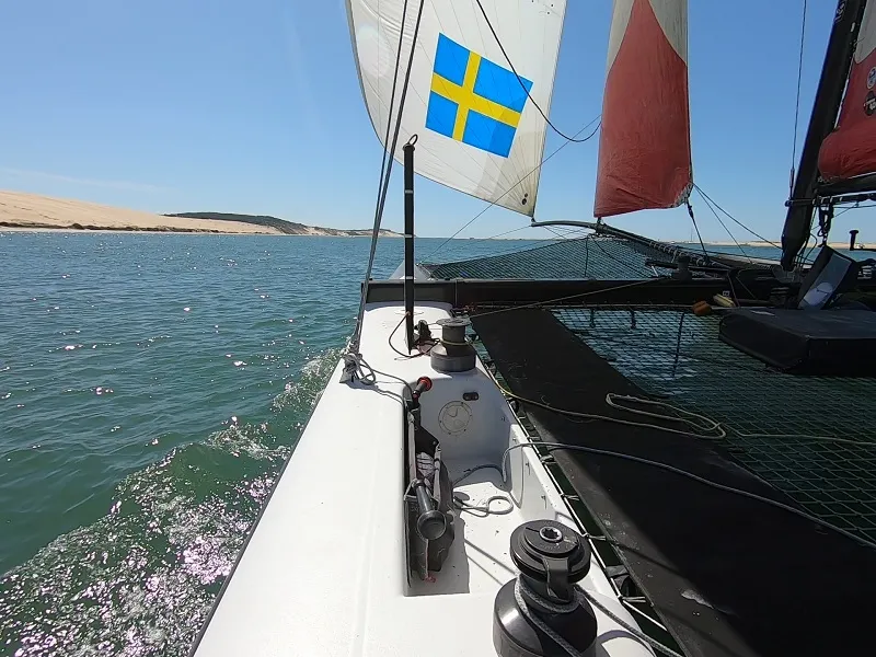 Sailing boat with Swedish flag sailing near Arcachon, with other sailing vessels visible in the background and sand dunes along the coastline.