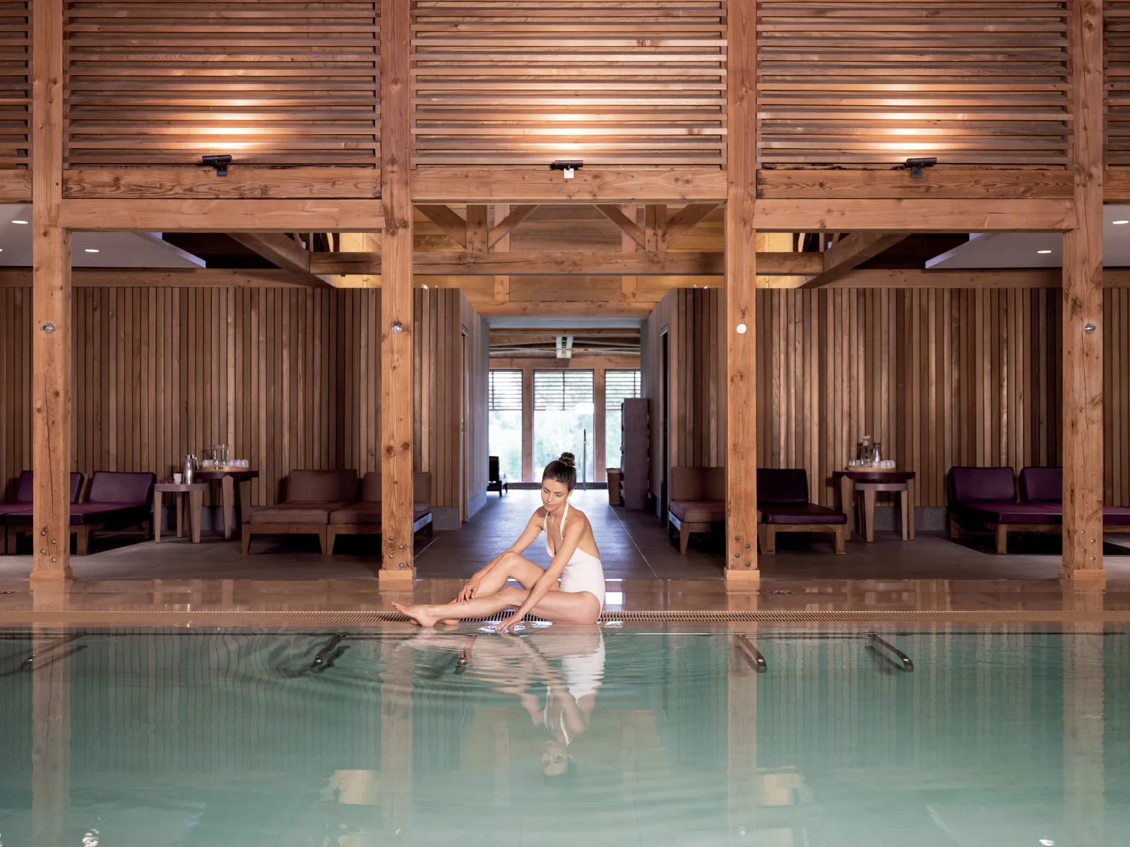 A woman sitting by the edge of an indoor swimming pool with turquoise waters in a spa with massive wooden structures, featuring relaxation cabins in the background at Sources de Caudalie in Bordeaux.