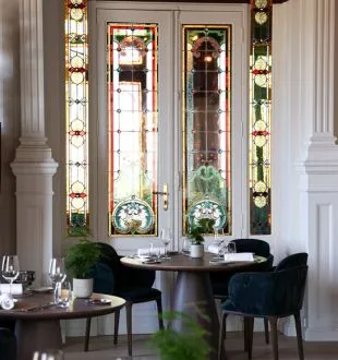 Elegant interior of Grand Barrail restaurant in Bordeaux featuring two black chairs facing tall glazed doors with golden patterns, decorated with Christmas garlands, white columns and green plants.