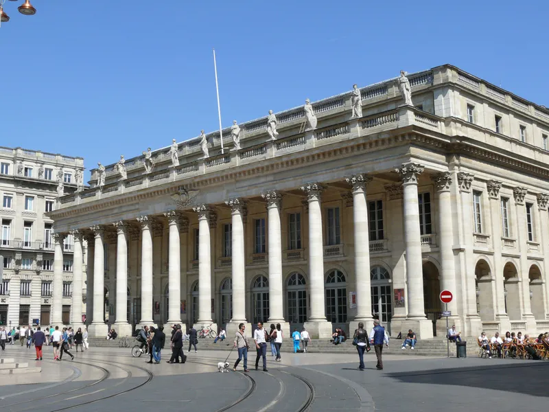 The Grand Theatre of Bordeaux featuring its neoclassical facade with white columns, topped with an ornate balustrade decorated with statues, under a clear blue sky with visitors in the square.