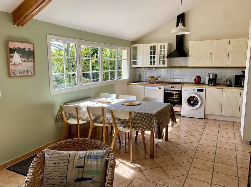 Bright spacious kitchen with pale green walls, large multi-pane windows, patterned tablecloth over dining table, natural wood chairs, white cabinetry, modern appliances and exposed ceiling beam