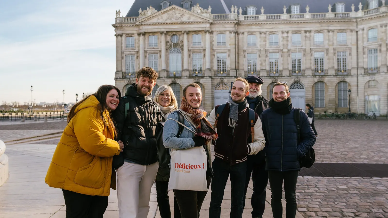 Seven smiling tourists posing in front of the Palais de la Bourse in Bordeaux, one holding a Délicieux! Food Tour bag.