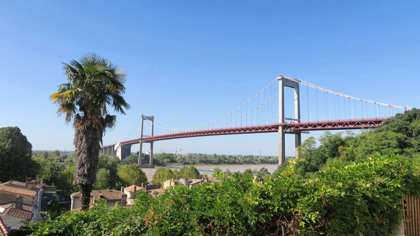 The Aquitaine Bridge spanning the Garonne River in Bordeaux, featuring its distinctive twin towers and suspension cables, surrounded by lush greenery and residential buildings.
