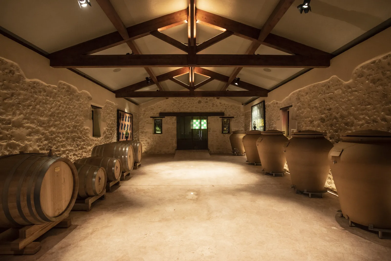 Wine cellar at Domaine des Perchés in Bordeaux featuring wooden barrels arranged under exposed timber beams and illuminated ceiling