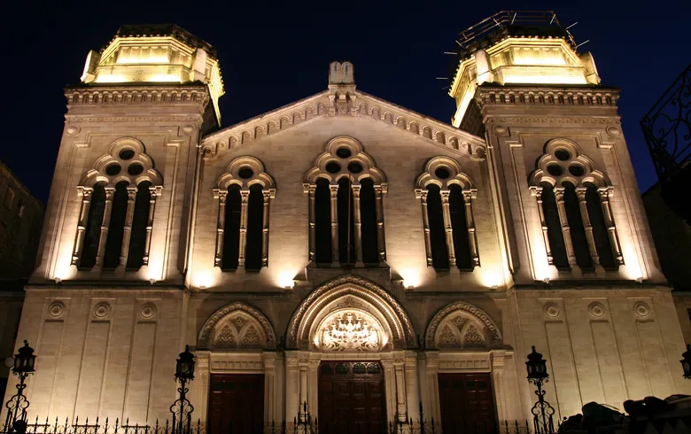 Night view of the illuminated façade of Bordeaux Synagogue, featuring Romano-Byzantine architecture with twin topped towers, paired lancet windows and three entrance porches.