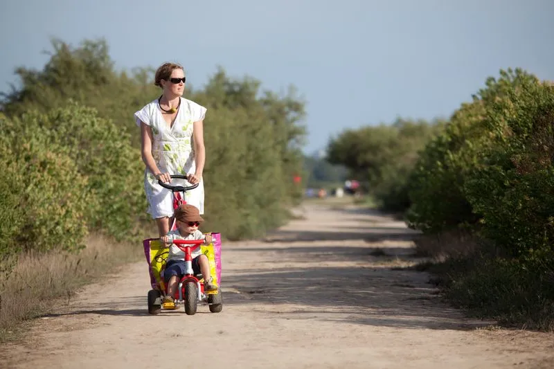A woman in a white dress and sunglasses pushes a pink and yellow tricycle on a dirt road lined with vegetation in the Certes-Graveyron estates near Bordeaux.