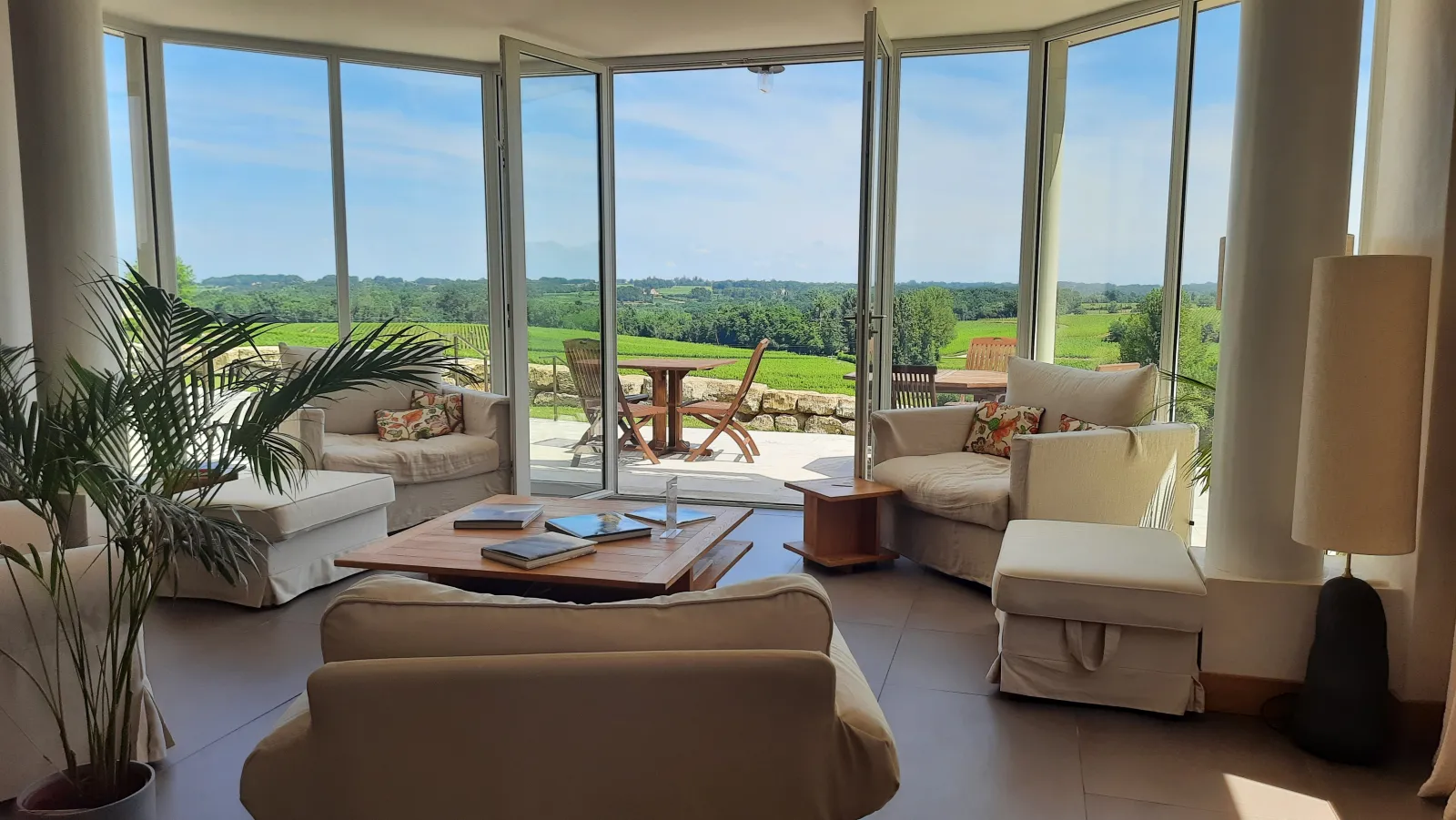 Spacious living room with panoramic glass doors overlooking Bordeaux vineyards, elegant beige furniture, green plants and wooden coffee table, outdoor terrace visible at Logis de Valandraud.