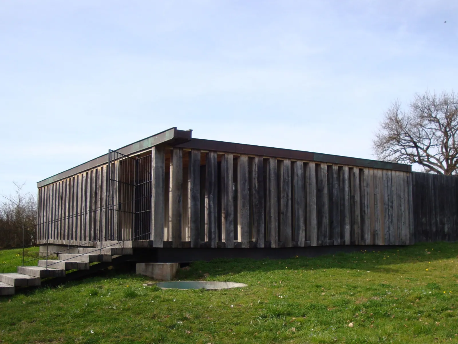Modern dark wooden building on stilts with access staircase, located in a grassy park with bare trees in the background, interpretation center structure for Pair-non-Pair Cave near Bordeaux