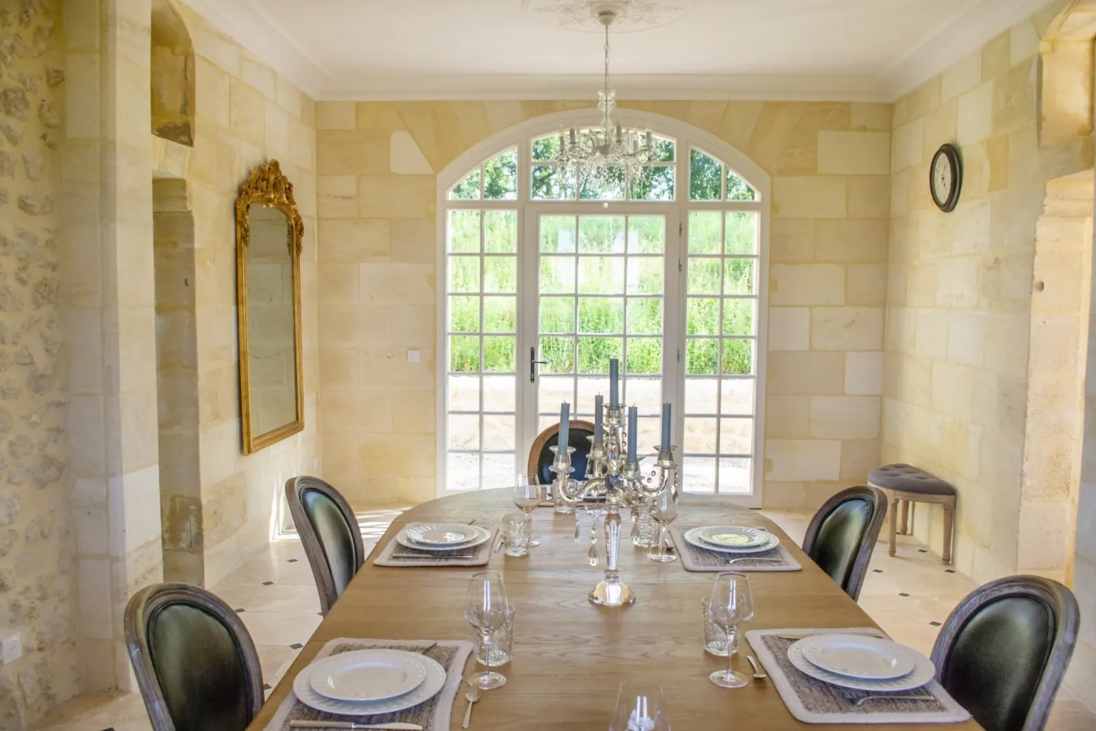 Elegant dining room at Château de Roques featuring a set table for four guests, black chairs, crystal chandelier, and arched French doors opening onto the green gardens.