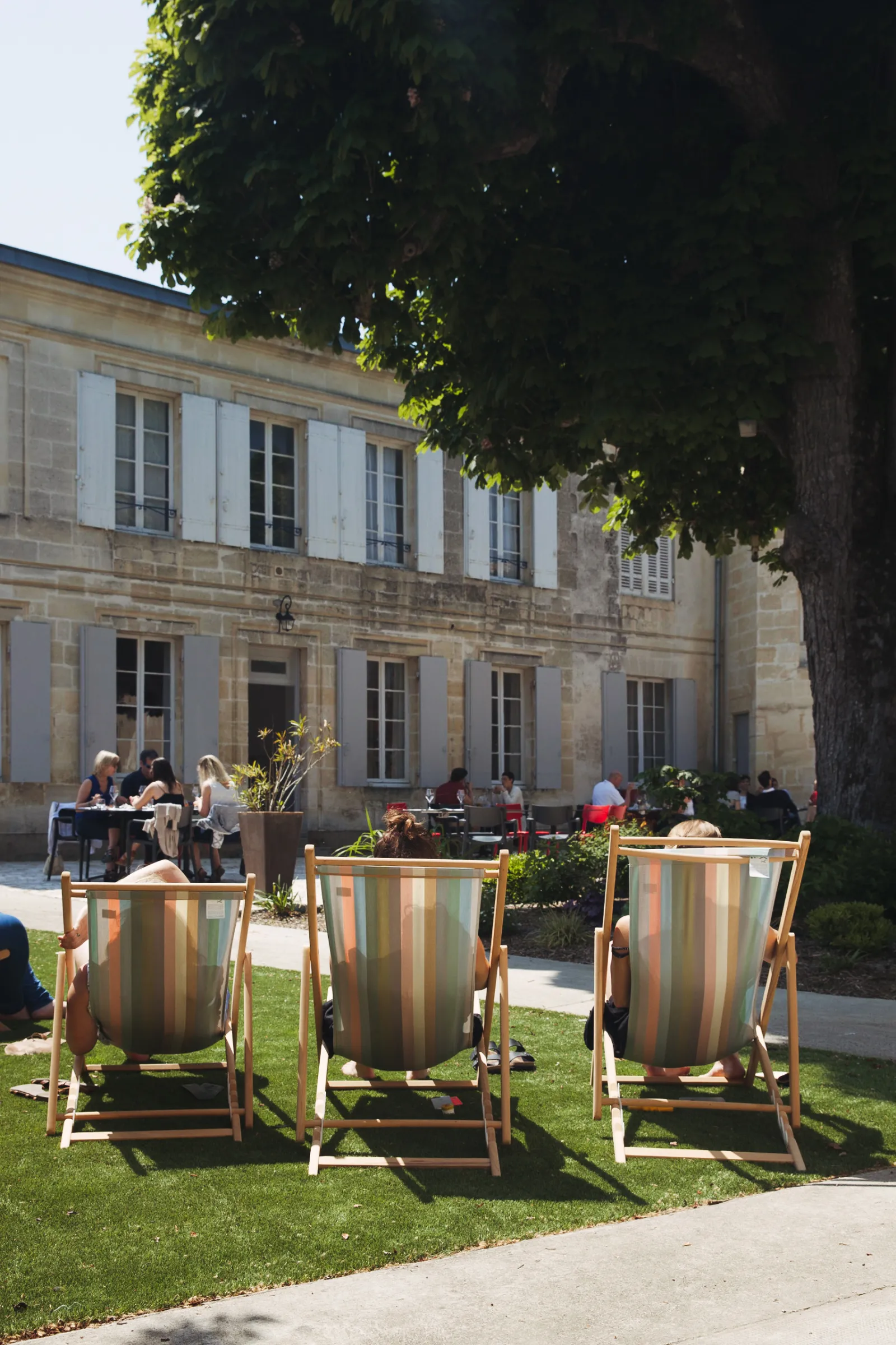 Three striped deckchairs on the lawn of Château Ambe Tour Pourret garden in Bordeaux, with visitors sitting in the background in front of the stone façade.