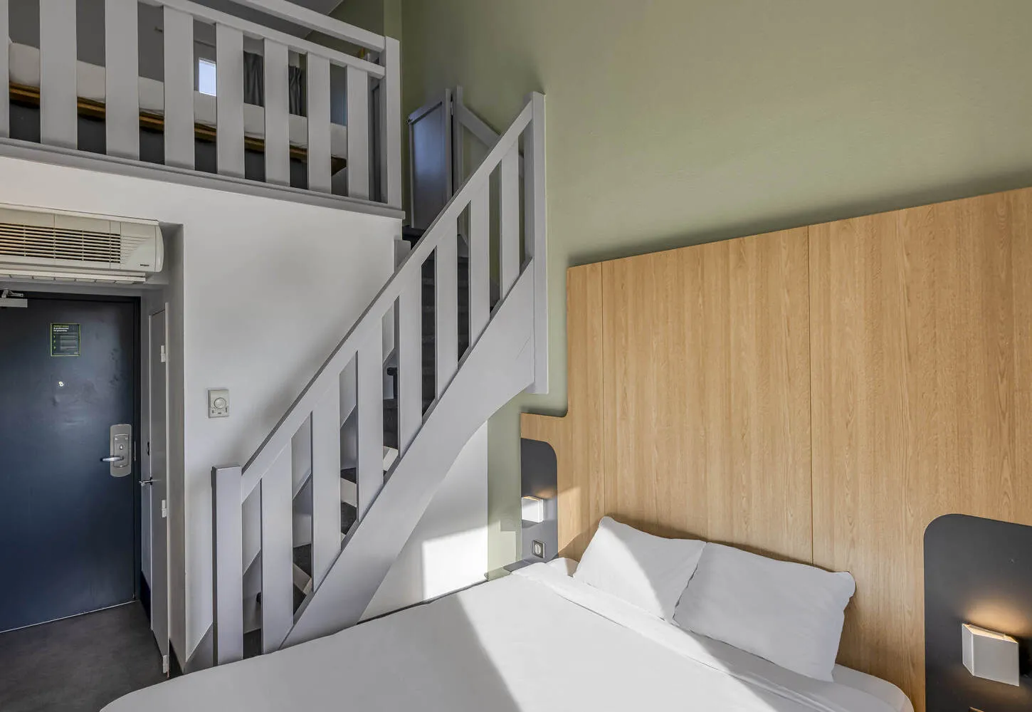 Modern hotel room featuring a double bed, white staircase leading to a mezzanine loft, light wood wall paneling, dark blue entrance door, and wall-mounted air conditioning unit
