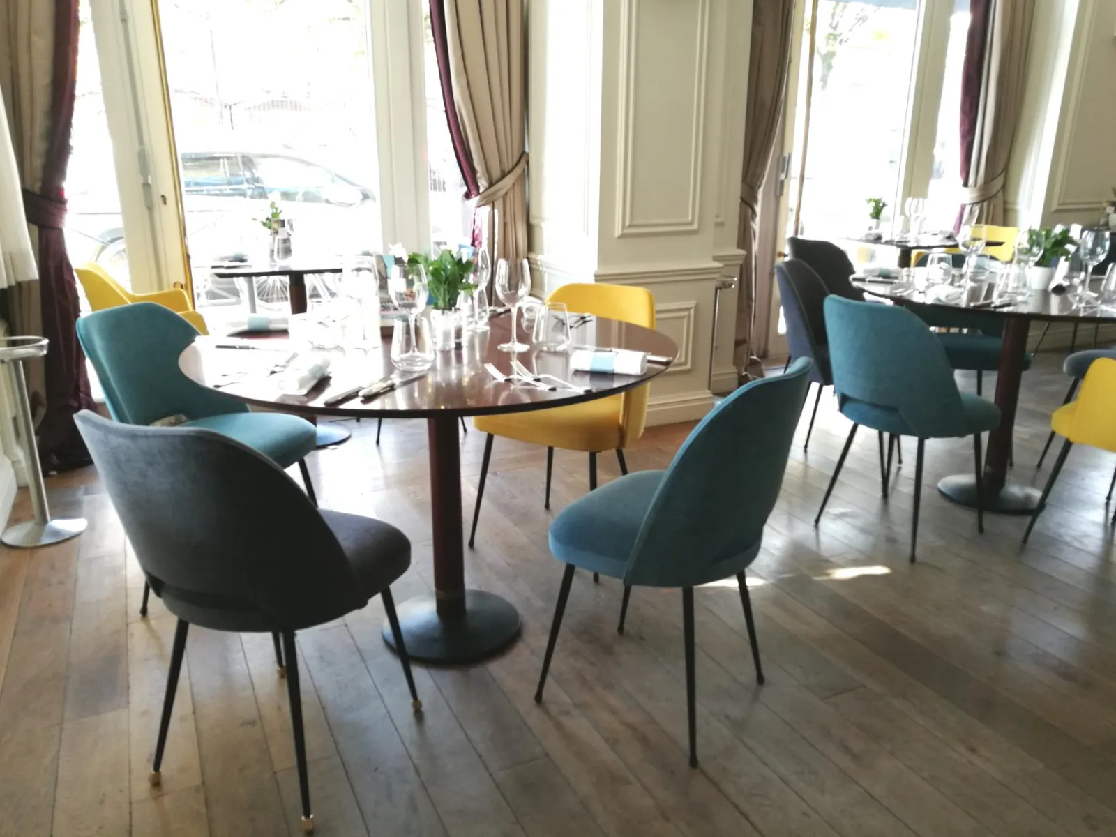 Elegant dining room at Comptoir de Sèze in Bordeaux featuring black round tables, blue and yellow upholstered chairs, large windows overlooking the street, floral arrangements and burgundy drapes.