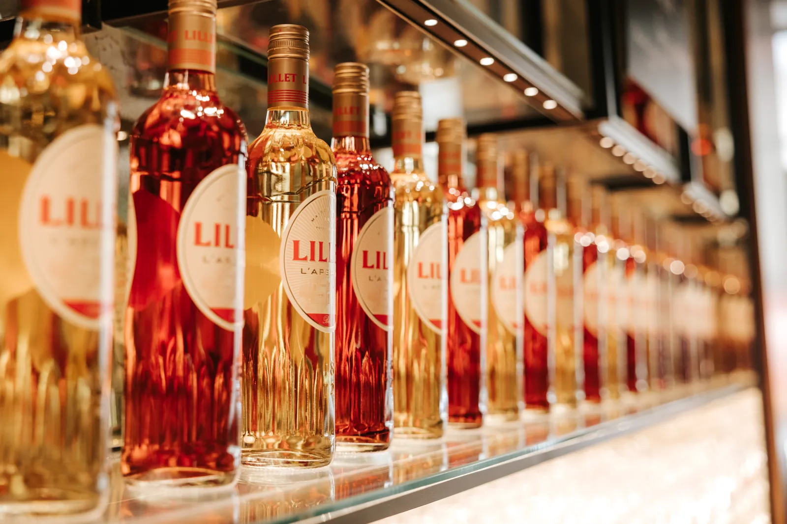 Row of Lillet bottles displayed on a shelf, featuring red and golden hues with their distinctive white labels clearly visible.