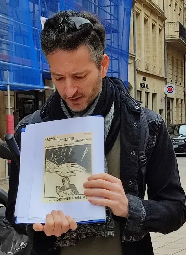 A man holds a historical French Passive Defense propaganda poster from World War II on a Bordeaux street.