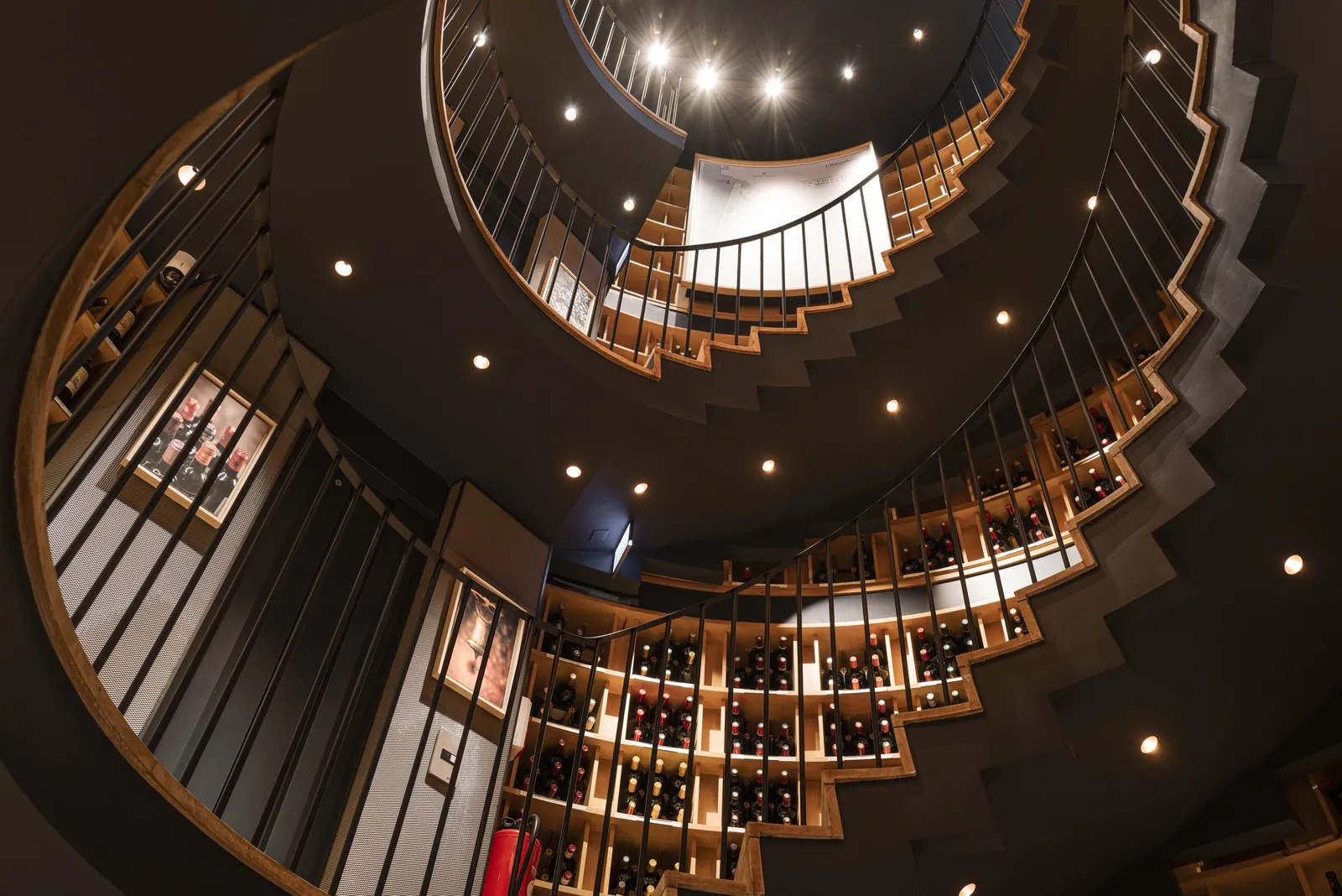 Overhead view of a circular wine cellar with wooden wine racks arranged in a spiral pattern, illuminated by spotlights, in the L'Intendant building in Bordeaux.