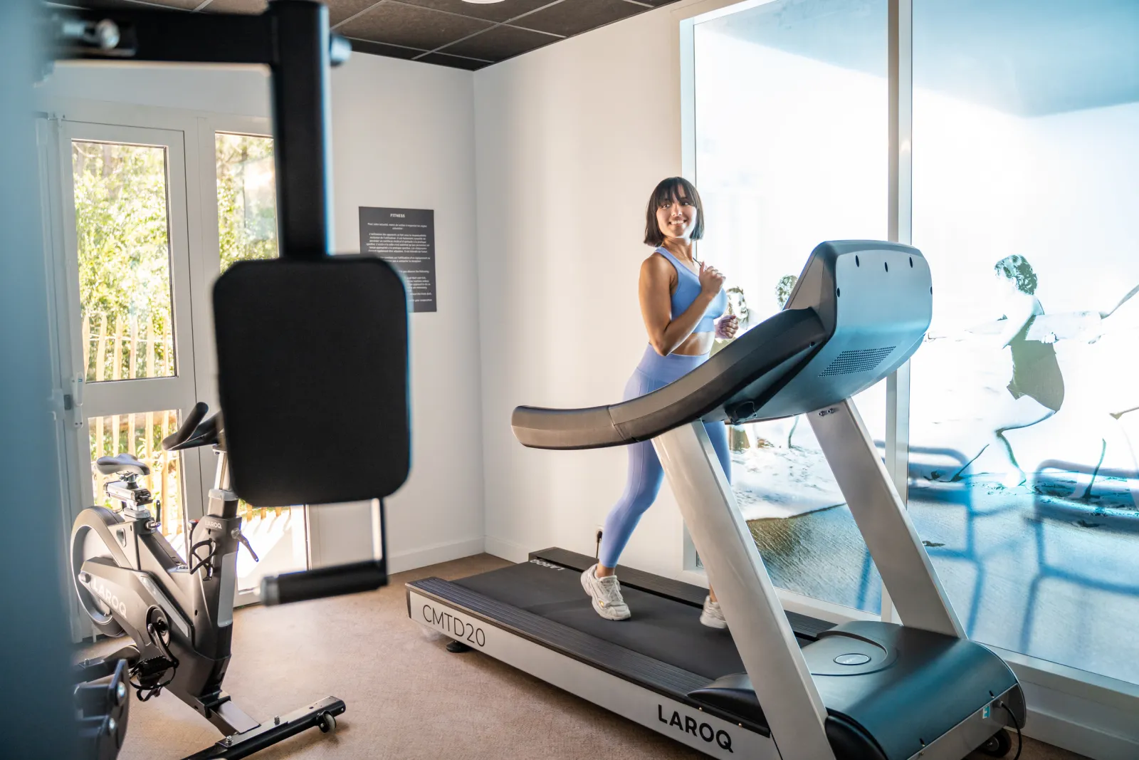 Woman in athletic wear running on a treadmill in a modern fitness room with large windows, with a stationary bike visible in the background